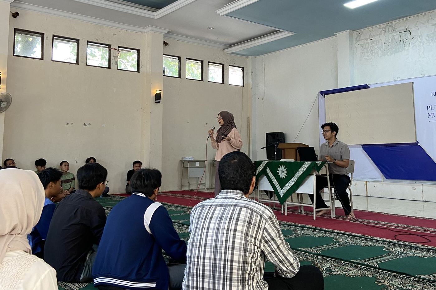 Sesi berbagi di aula berkarpet hijau, pembicara memegang mikrofon di depan peserta